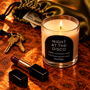 A lit scented candle in a clear glass jar sits on a patterned copper-toned surface, surrounded by lifestyle props including a lipstick, keys, cash, and a gold satin drawstring bag. The candle label reads 'NIGHT AT THE DISCO' with scent text 'AMBER, MAHOGANY, MUSA'.