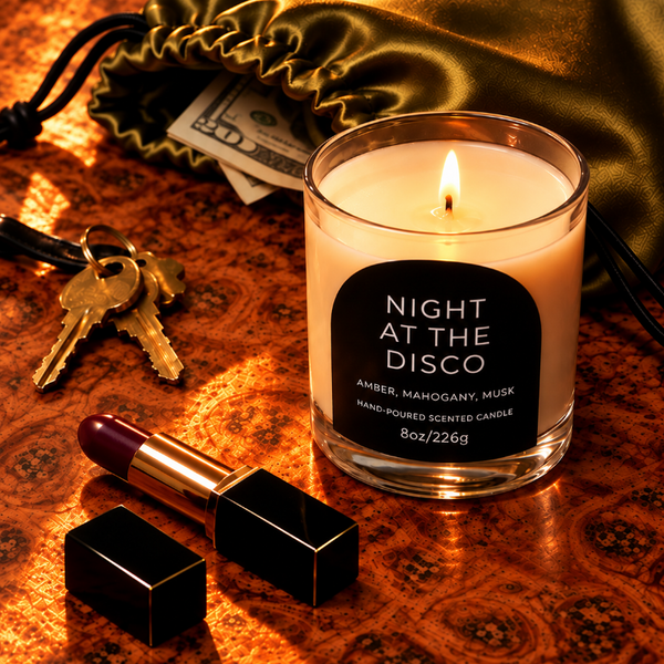 A lit scented candle in a clear glass jar sits on a patterned copper-toned surface, surrounded by lifestyle props including a lipstick, keys, cash, and a gold satin drawstring bag. The candle label reads 'NIGHT AT THE DISCO' with scent text 'AMBER, MAHOGANY, MUSA'.